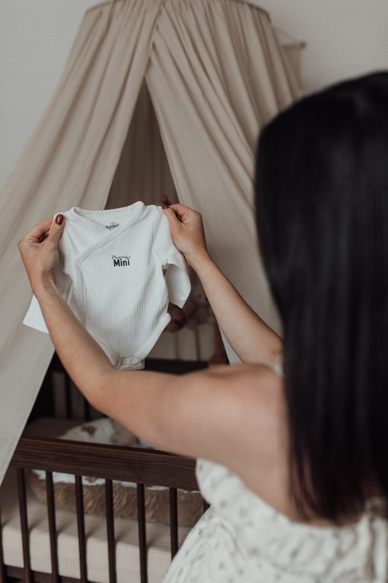 Werdende Mutter hält einen Babybody vor dem Kinderbett